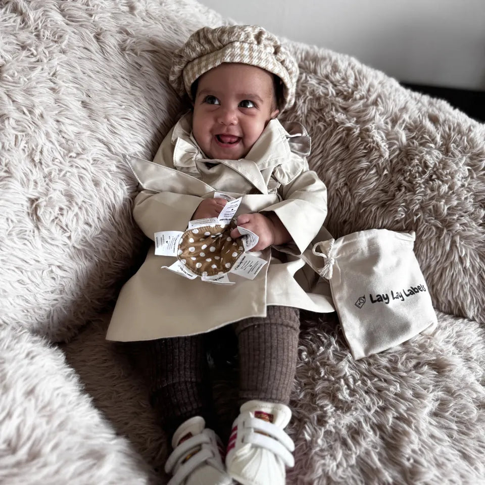 A smiling baby in a cap and trench coat holding a Polka Dot Lay Lay Mini, with the cotton gift bag beside her