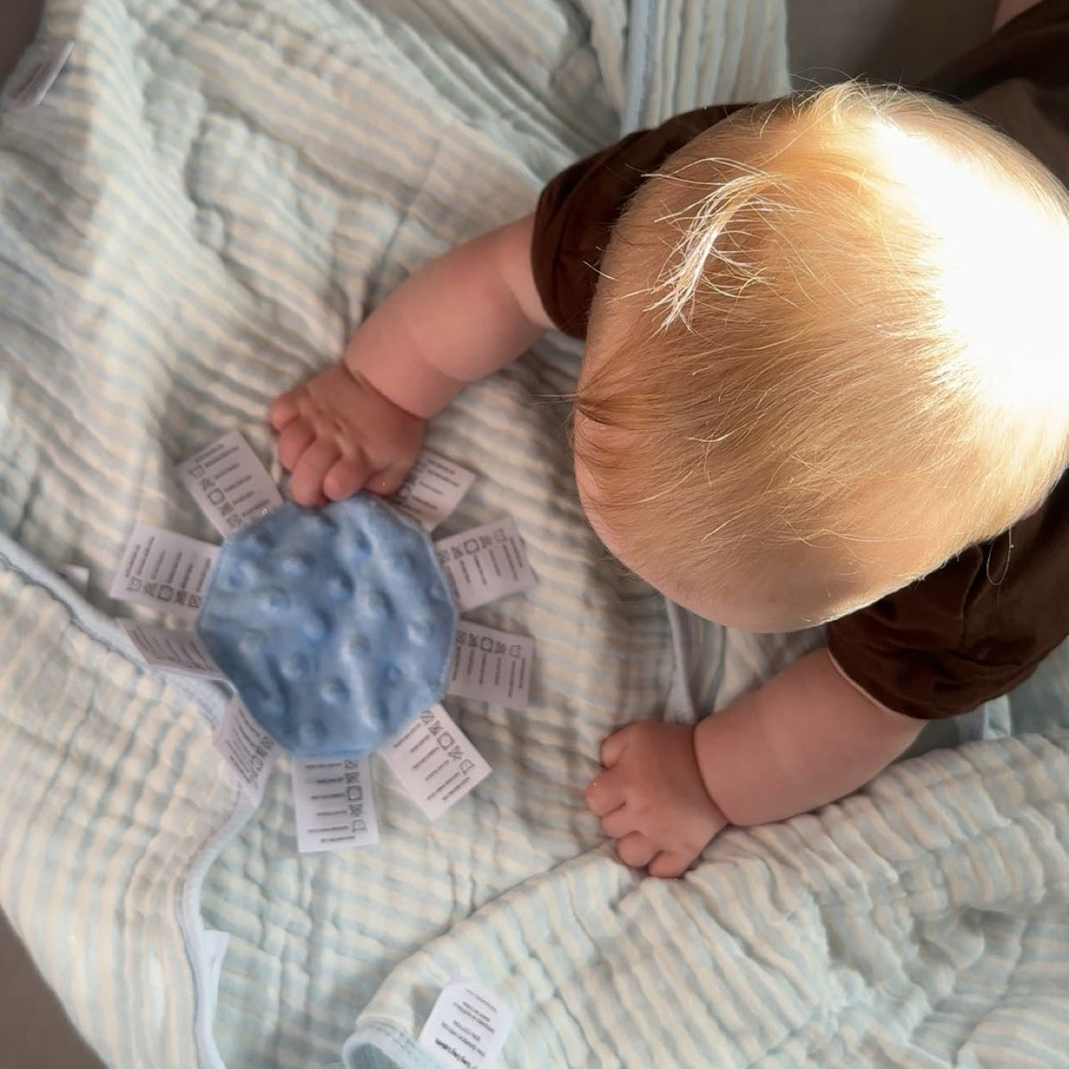 Baby holding blue sensory mini with labels while lying on soft blanket